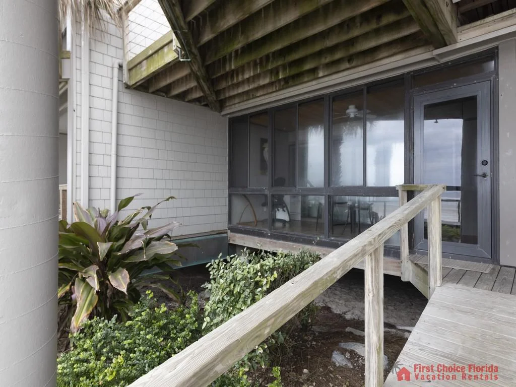 Wooden walkway leads to a screened balcony with ocean views, surrounded by gray siding and green landscaping.