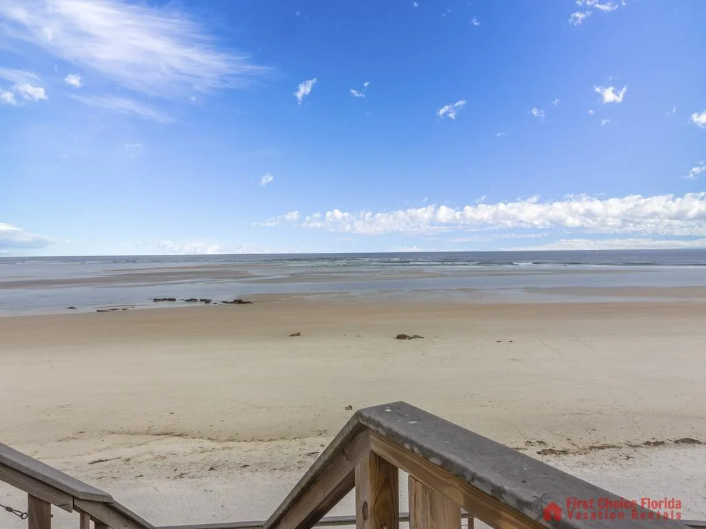 View over the wooden staircase railing to a wide sandy beach and calm ocean under a blue sky with scattered clouds.