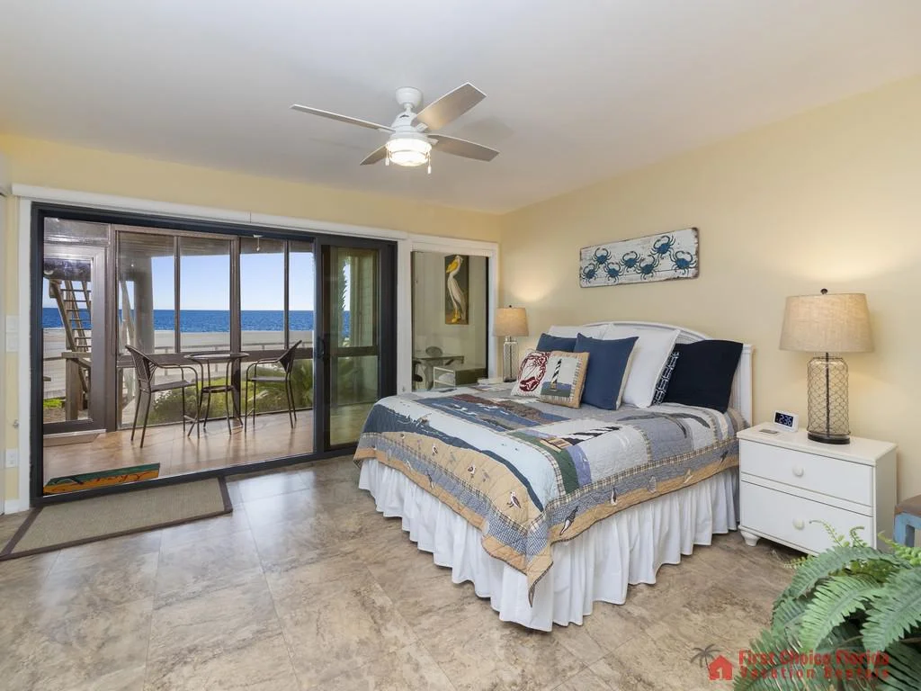 Master bedroom with tiled floors, coastal-themed bedding, glass sliding doors opening to an oceanfront balcony.