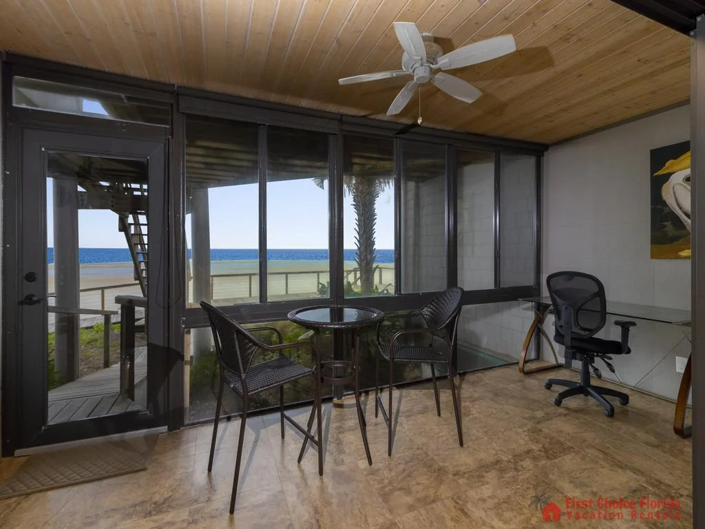 Enclosed balcony with wicker bar chairs and table, glass door opening to beachside walkway and ocean views.
