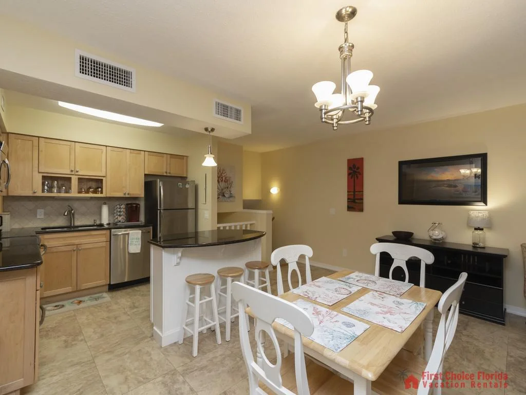 Wood dining table with white chairs and coastal-themed placemats sits beside a kitchen bar with three wooden stools.