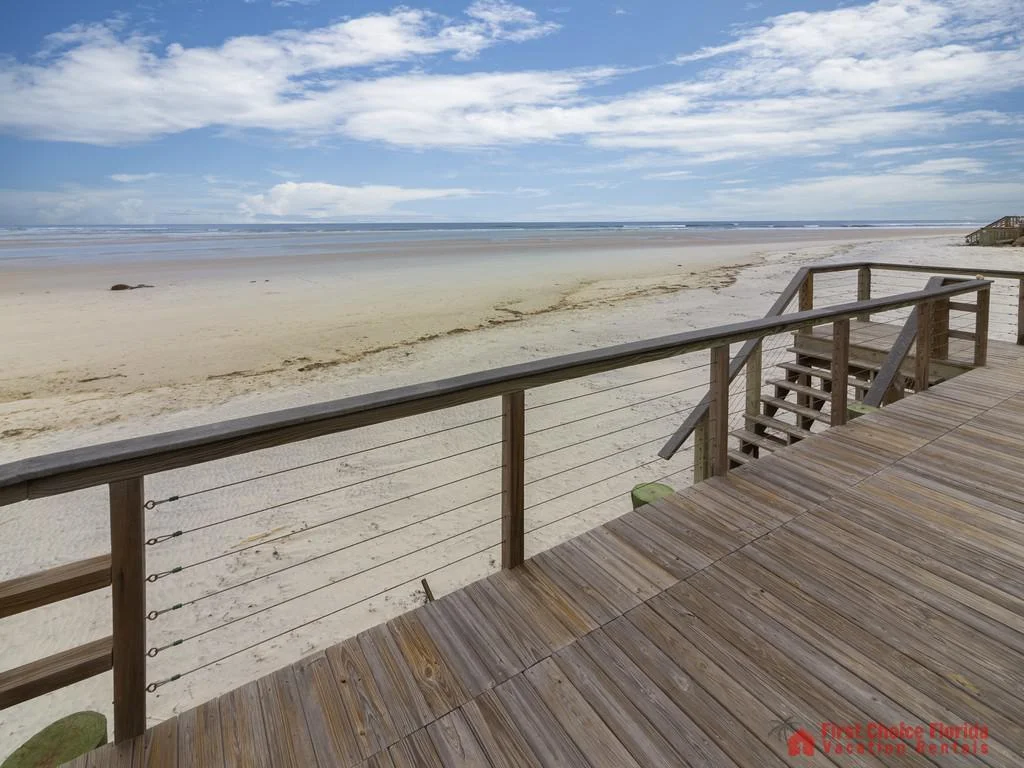 Wooden balcony with cable railing overlooks wide sandy beach and calm ocean waves under a partly cloudy sky.