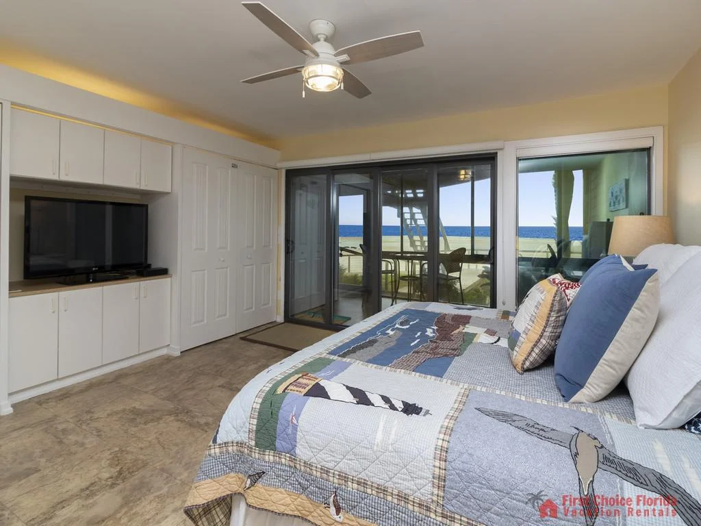 Master bedroom features a queen bed with nautical quilt, ceiling fan, and sliding glass doors opening to ocean views.