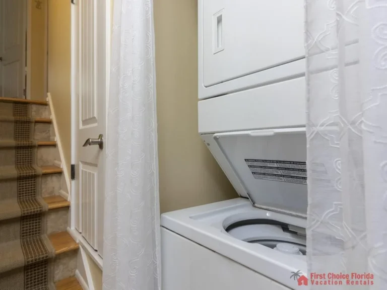 Stacked white washer and dryer sit next to carpeted stairs with a patterned runner and a white door.