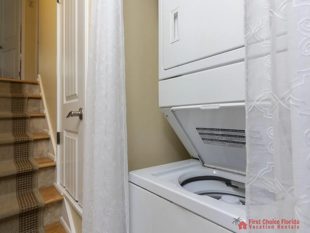 Stacked white washer and dryer sit next to carpeted stairs with a patterned runner and a white door.