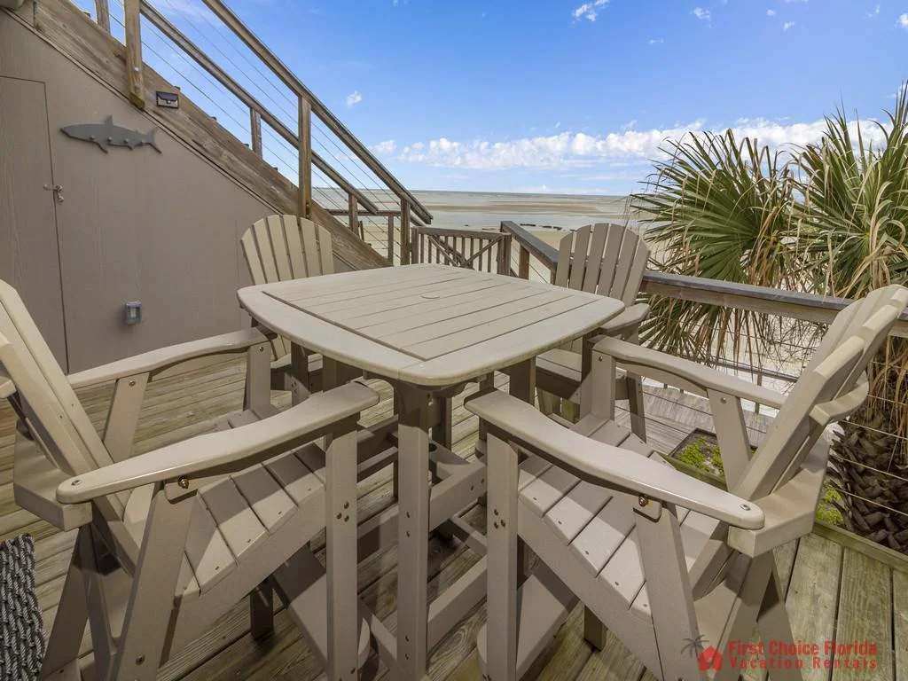 Balcony deck with tan Adirondack chairs and square table overlooks sandy beach and ocean horizon.