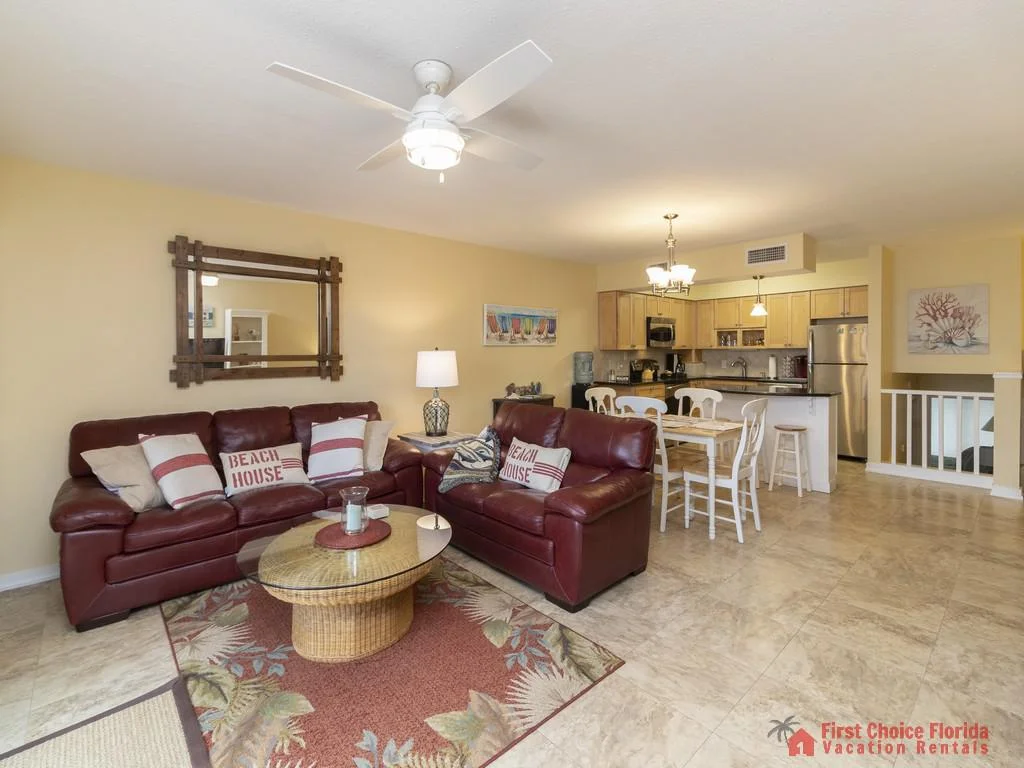 Living area with maroon leather sofas, wicker coffee table on a floral rug, and a kitchen with light wood cabinetry.