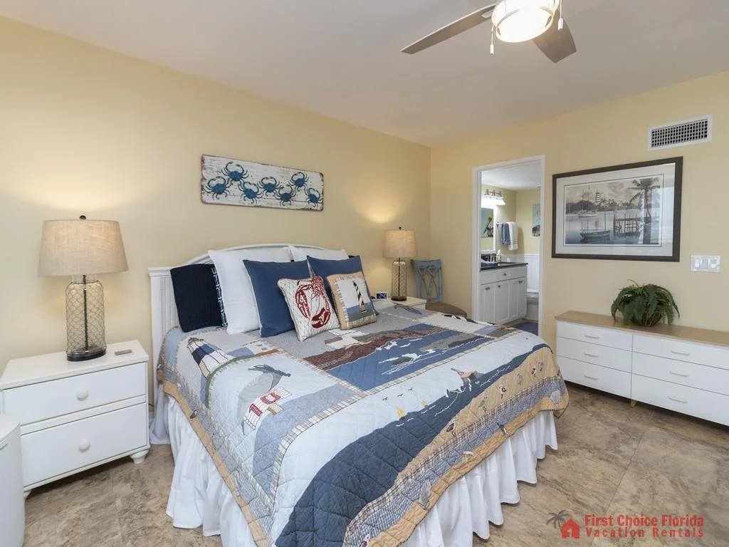 Bedroom with nautical-themed quilt, blue accent pillows, white nightstands, and a dresser beneath framed artwork.