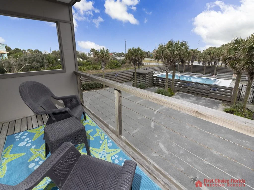 Balcony with dark wicker chairs and blue starfish-patterned rug overlooks fenced communal pool and palm trees.