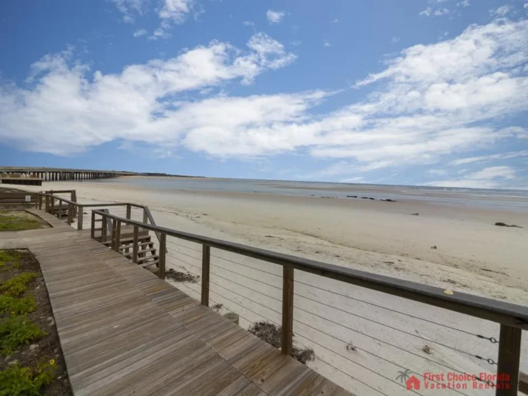 Wooden boardwalk with cable railing leads down to a wide sandy beach under a partly cloudy sky.