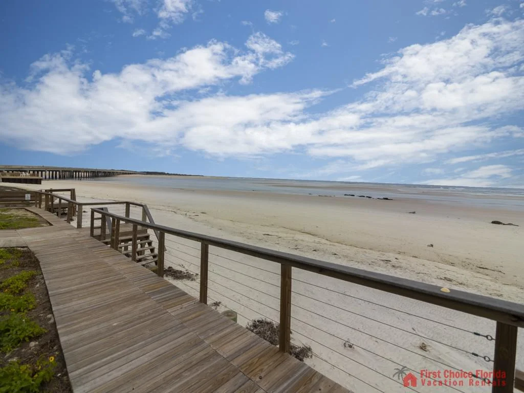 Wooden boardwalk with cable railing leads down to a wide sandy beach under a partly cloudy sky.