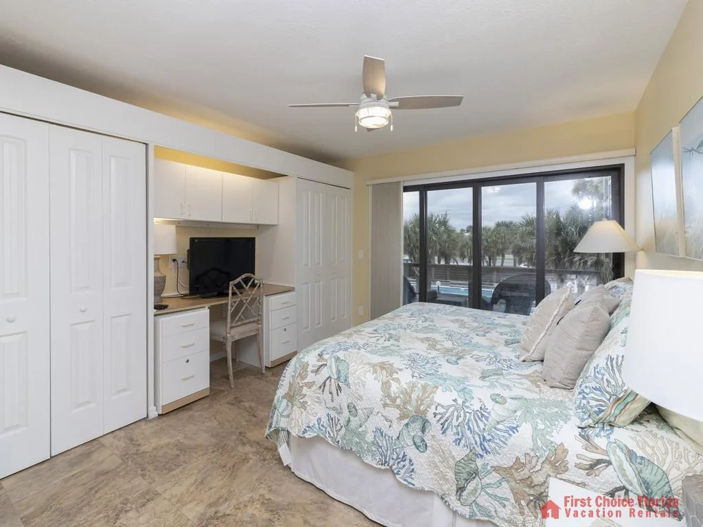 Bedroom with coastal-patterned bedding, built-in white cabinetry desk, and sliding glass doors opening to a poolside balcony.