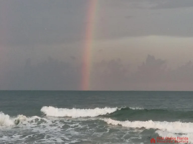 Ocean waves roll toward shore beneath a cloudy sky with a faint rainbow emerging in the distance.