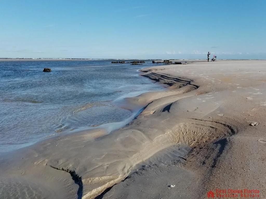 Tidal pools and wet sand patterns stretch along the shoreline under a clear blue sky with distant beachgoers.