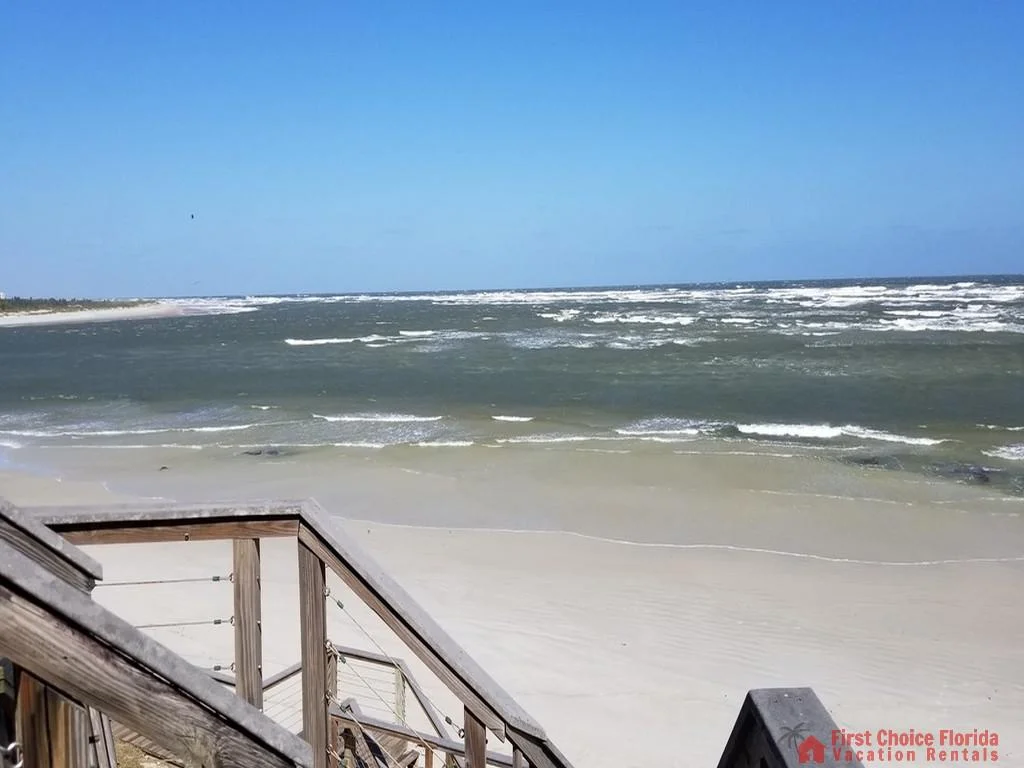 Weathered wood railing and steps lead down to a wide sandy beach with gentle ocean waves under a clear blue sky.