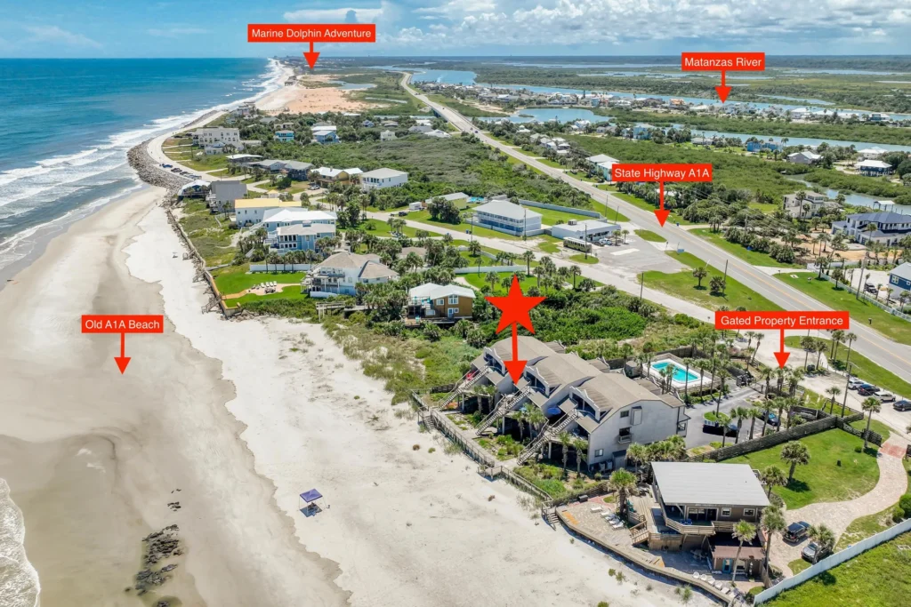 Gated entrance with black metal fencing, palm trees, and a paved driveway leading to beachfront condos.