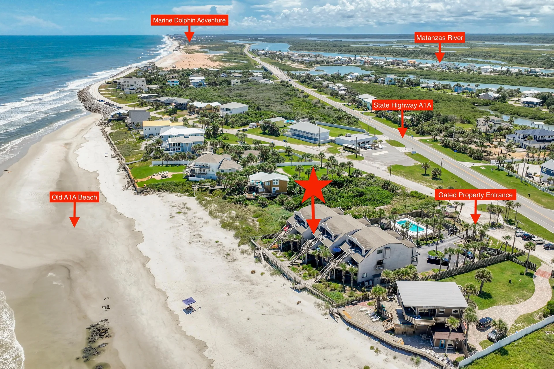 Gated entrance with black metal fencing, palm trees, and a paved driveway leading to beachfront condos.