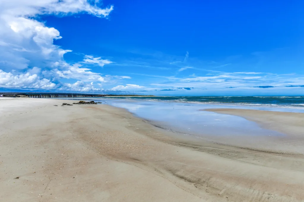 Wide sandy beach with shallow tidal pools stretches toward a long pier beneath a bright blue sky.
