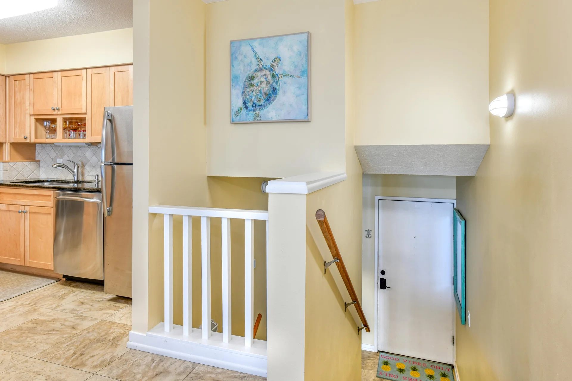 Entryway with white door and wooden handrail, adjacent to kitchen featuring maple cabinets and stainless steel appliances.