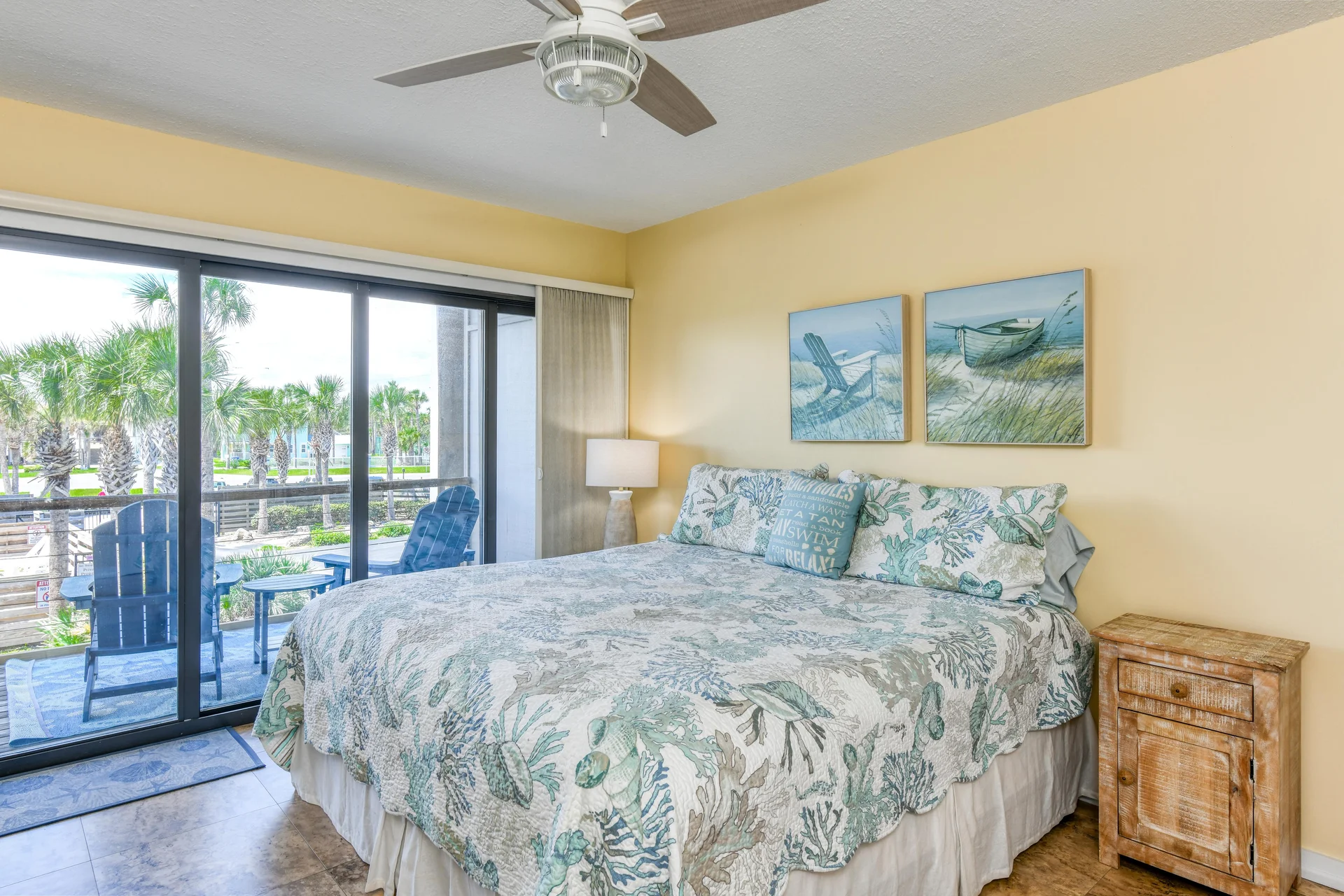 King bed in bedroom with coastal-patterned bedding, rustic nightstand, and sliding doors opening to a balcony with blue Adirondack chairs.