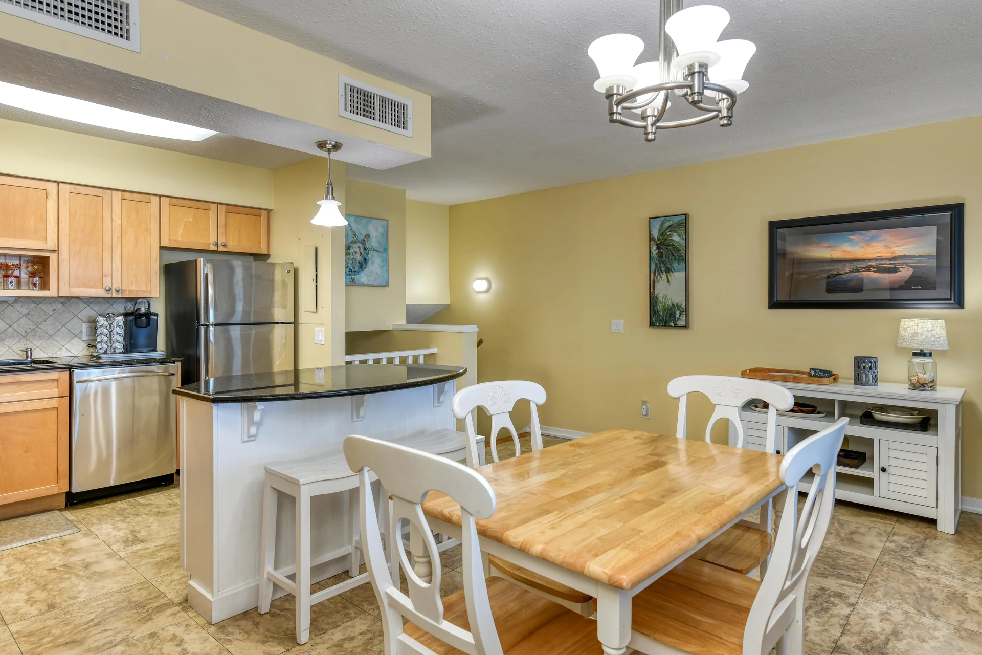 Dining area features a light wood table with white chairs beneath a brushed nickel chandelier and adjacent kitchen bar.