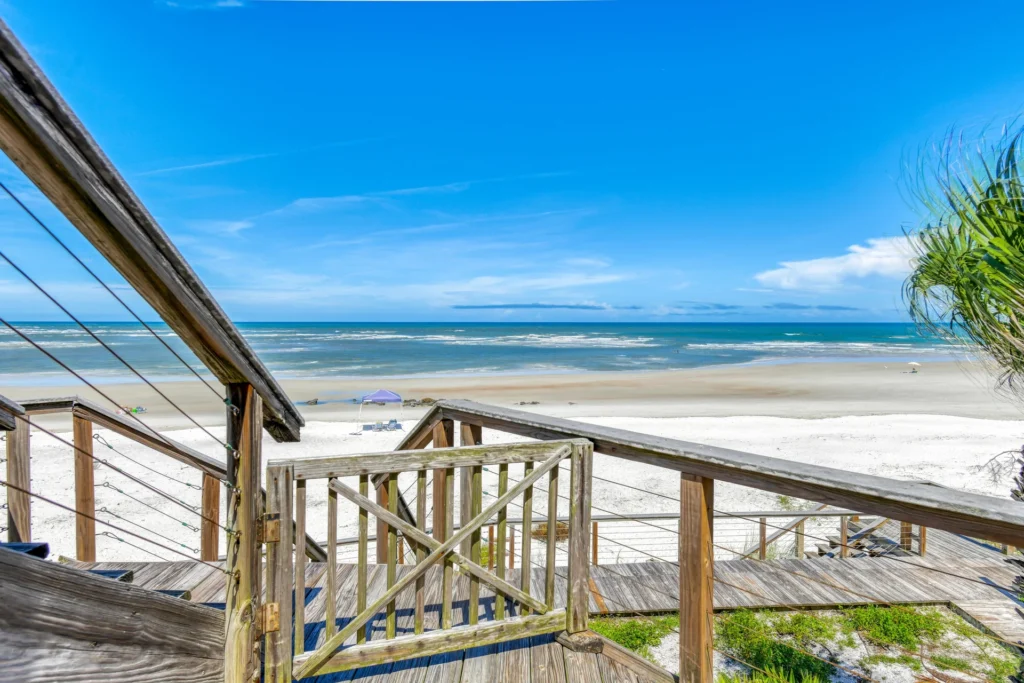 Weathered wooden gate opens to a stairway with cable railings descending toward the sandy beach and ocean waves.
