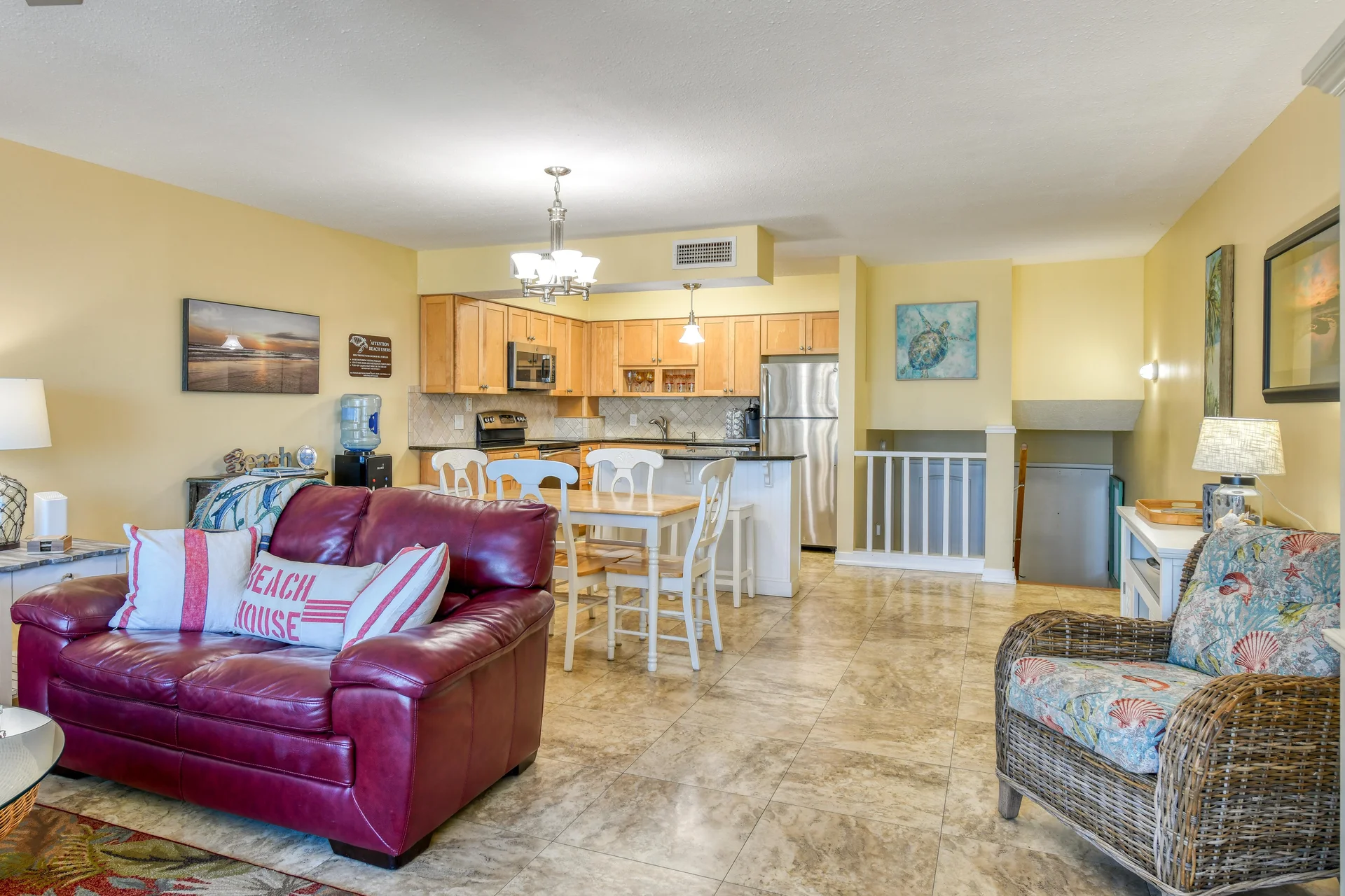 Living area with a burgundy leather loveseat, wicker armchair with seashell fabric, and tiled floors flowing into the kitchen.