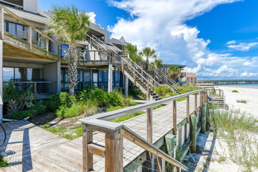 Boardwalk with wooden railing runs alongside beachfront condos with metal cable accents and palm trees nearby.