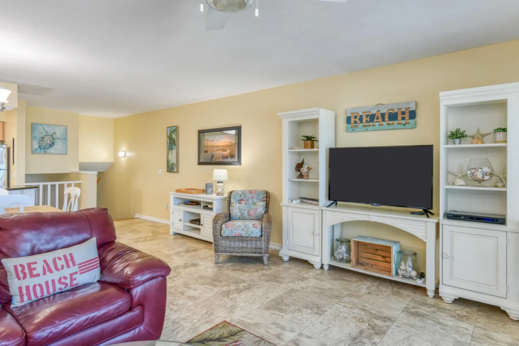Living room features a burgundy leather sofa with a "Beach House" pillow, wicker armchair, and white entertainment center.
