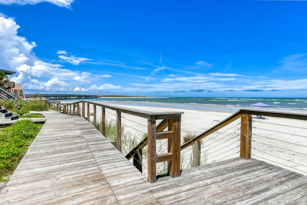 Wooden pathway with cable railings curves alongside dune grasses, leading to stairs that descend onto a white sandy beach.