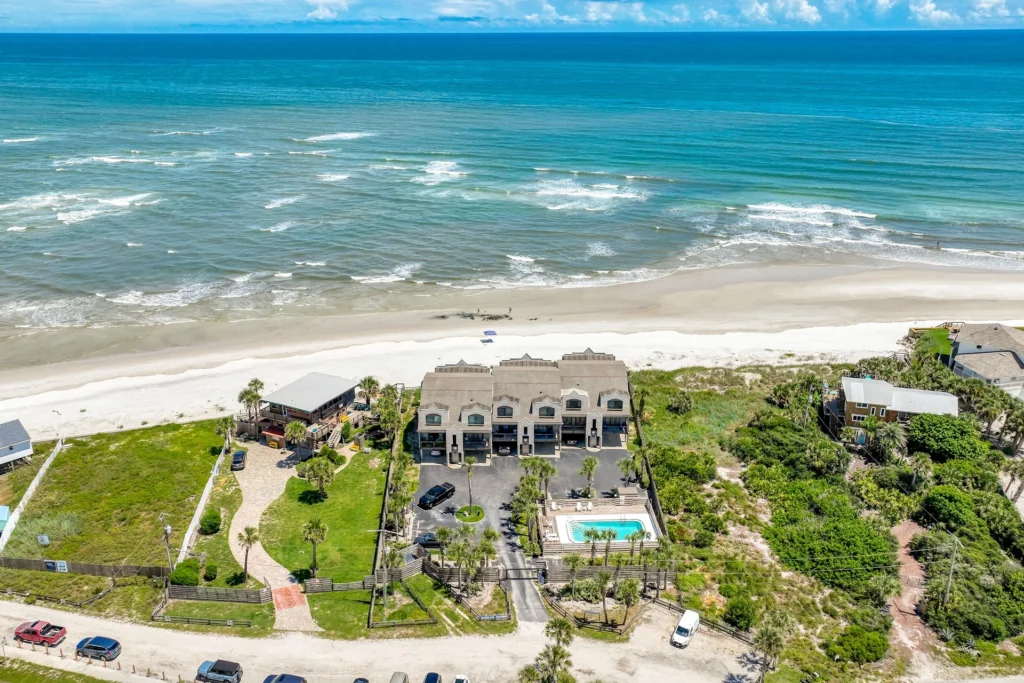 View from above showing the condo’s private parking, fenced pool area with palm trees, and sandy beach access nearby.