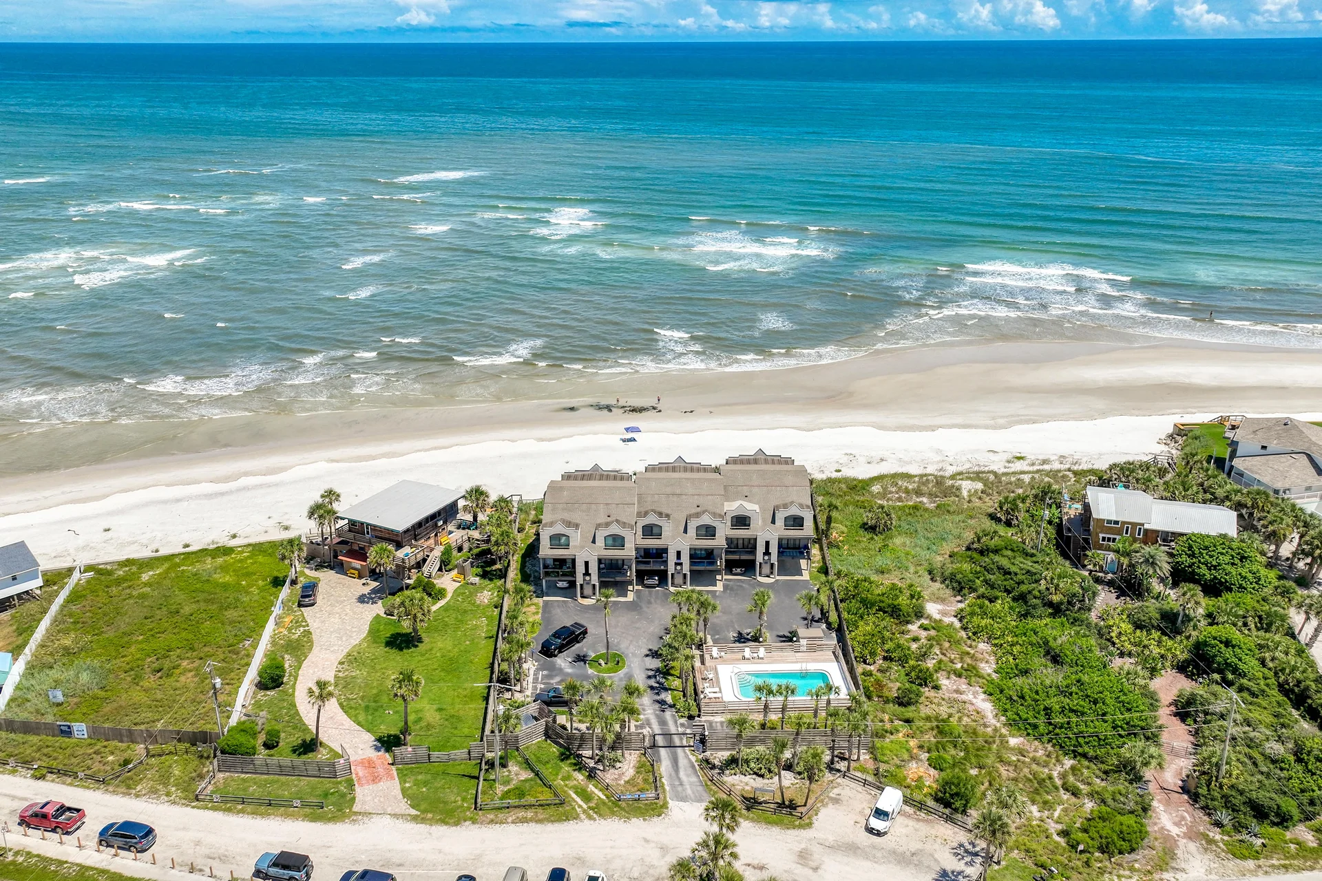 View from above showing the condo’s private parking, fenced pool area with palm trees, and sandy beach access nearby.