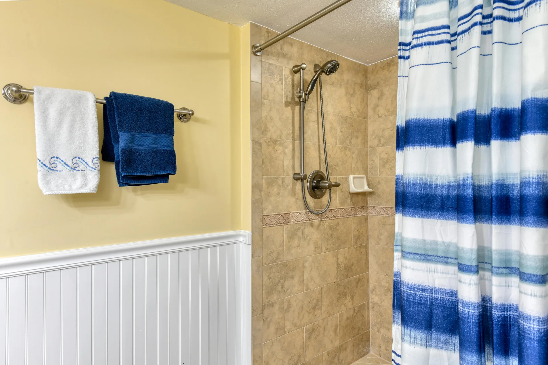 Walk-in shower features beige tile walls with decorative trim, brushed nickel fixtures, and a blue-striped shower curtain.