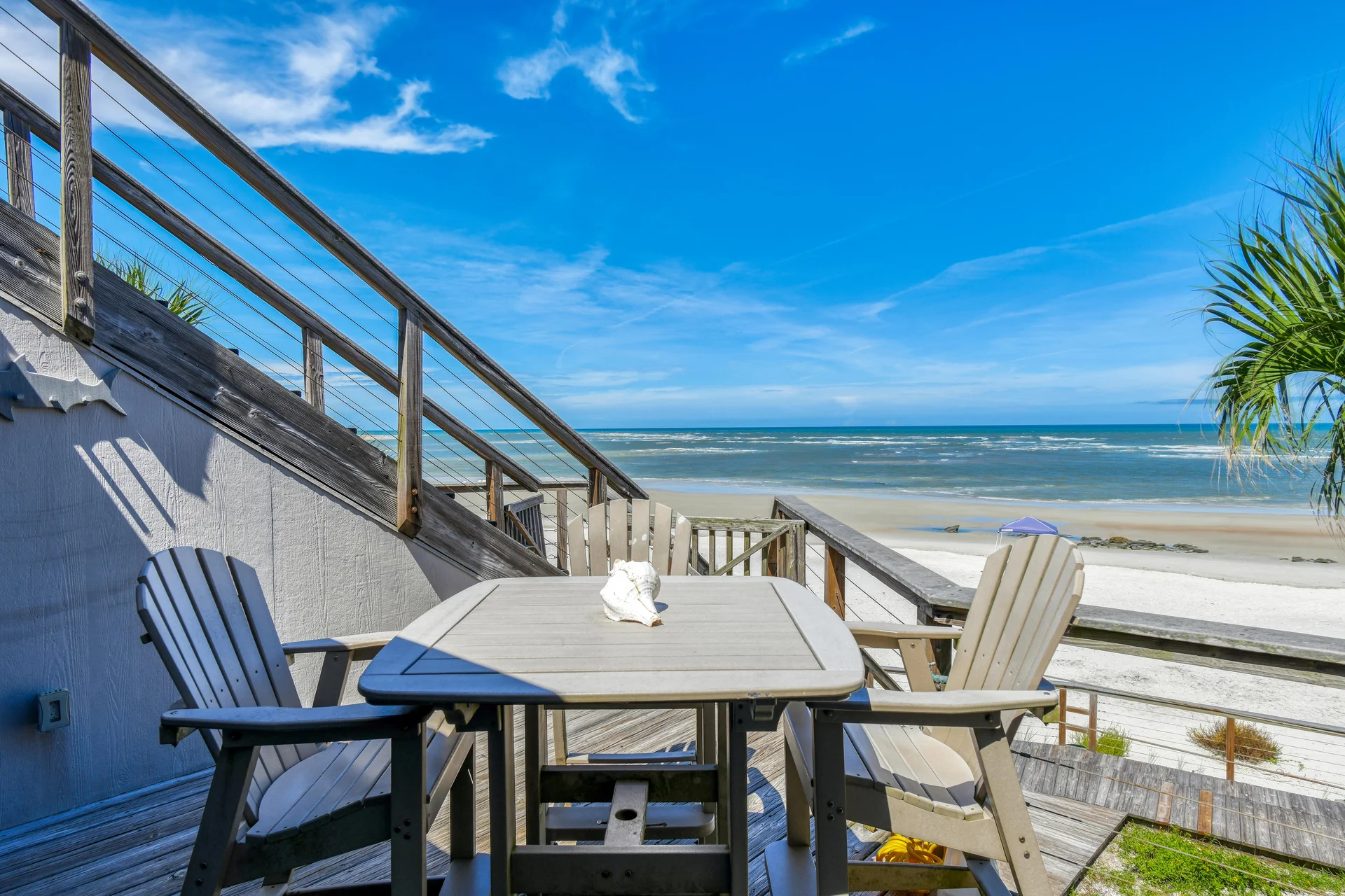 Square wooden outdoor table with a conch shell centerpiece surrounded by four beige Adirondack chairs on the deck.