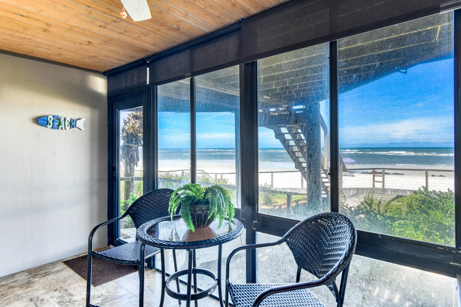Screened porch with wicker chairs, a glass-top table, a potted fern, and ocean views through large windows.