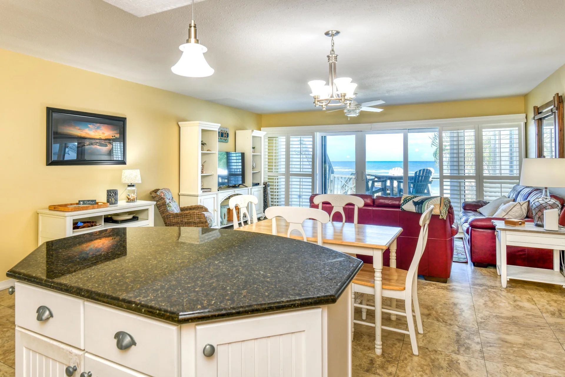 Kitchen island with dark granite countertop overlooks white dining set and red leather sofas facing ocean-view sliding doors.