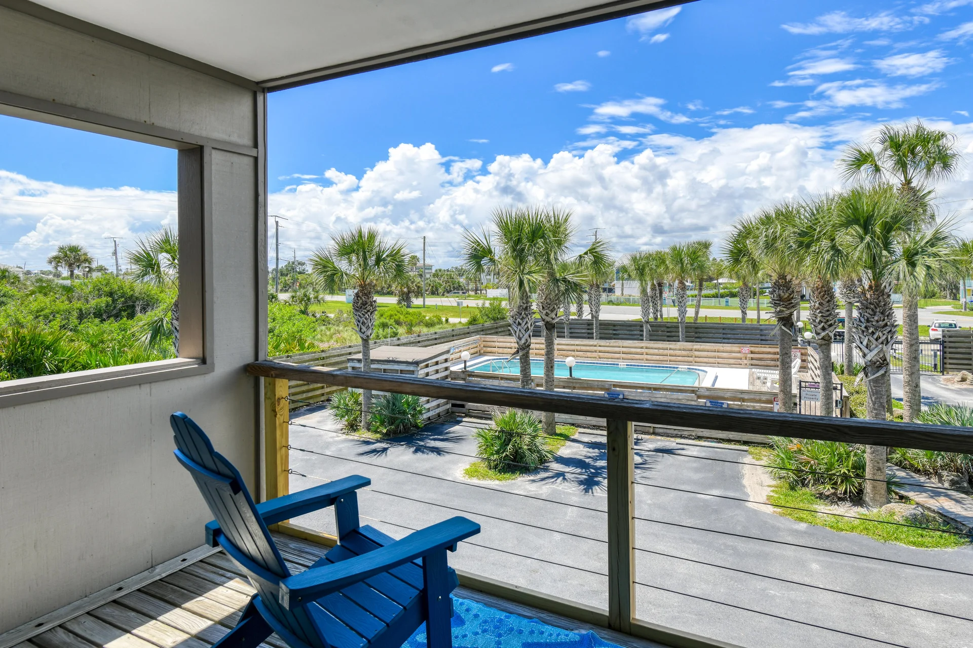 Covered balcony with a blue Adirondack chair overlooks a fenced pool area surrounded by palm trees.
