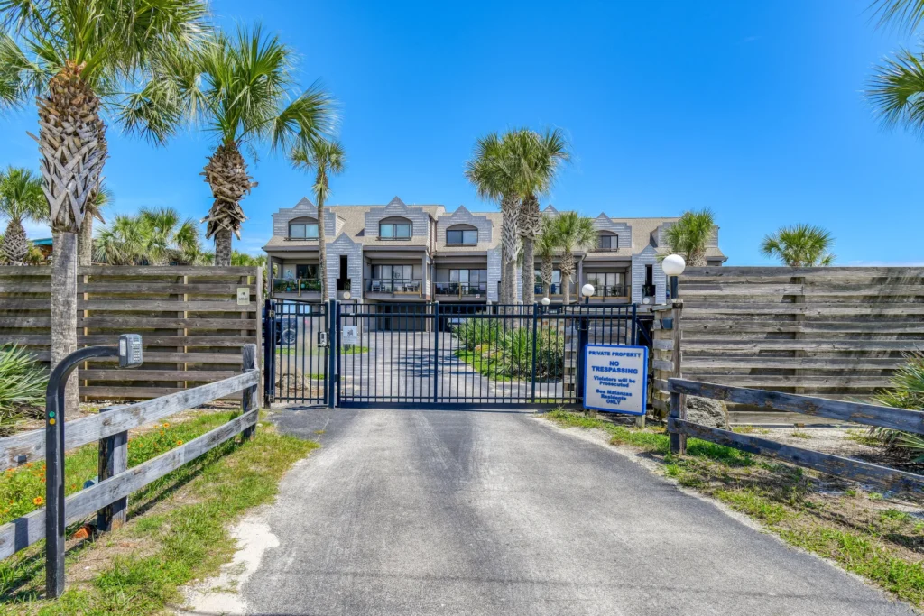 Black metal gate secured with keypad sits at entrance to beachfront condo complex, flanked by palm trees and weathered wood fencing.