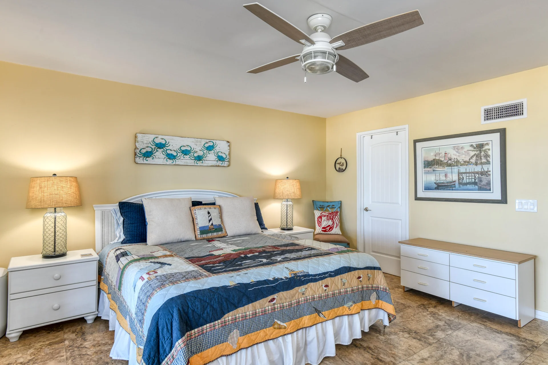 King bedroom with yellow walls, coastal-themed quilt and pillows, white nightstands, and ceiling fan.