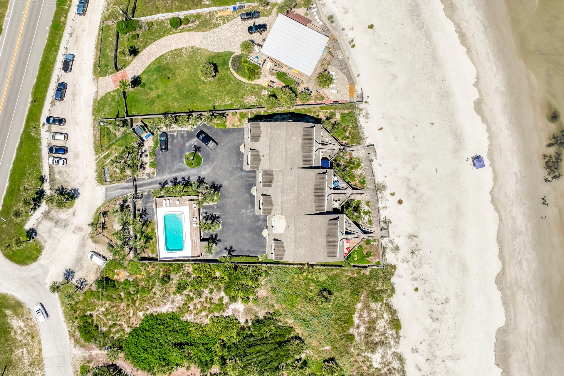 Overhead view shows beachfront condos, a fenced pool area, parking lot, and wide sandy beach meeting calm ocean waters.