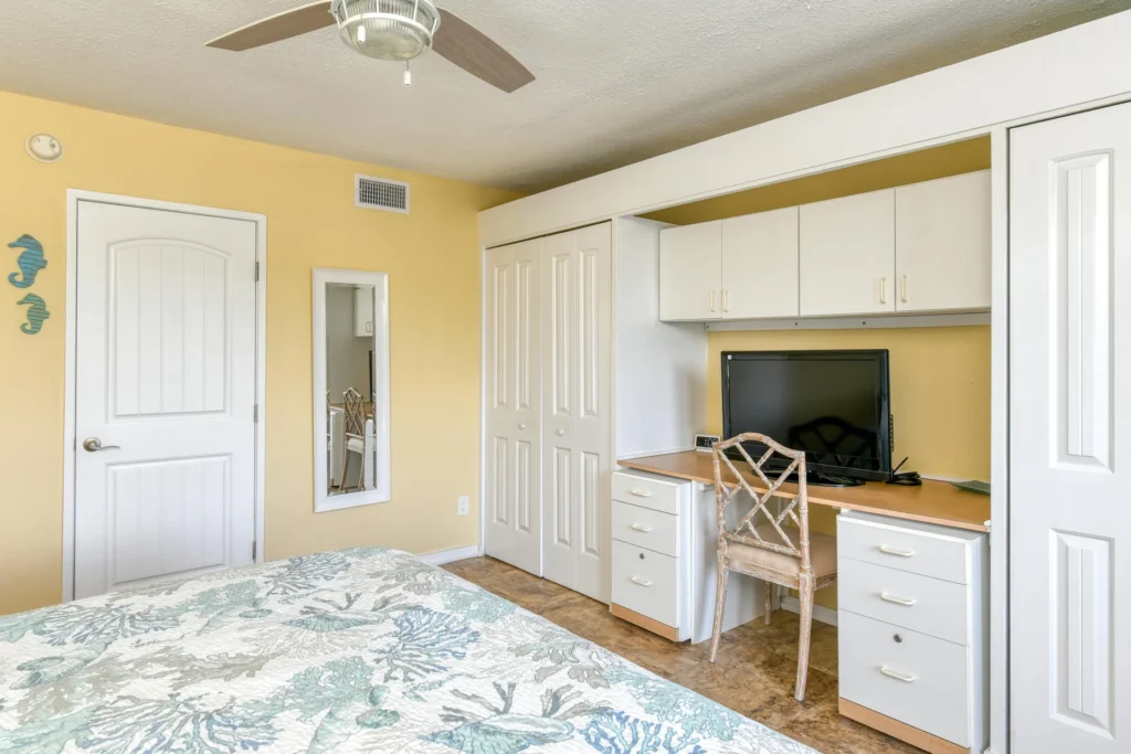 Built-in desk with white drawers and cabinets, a cushioned wooden chair, and a wall-mounted mirror in the bedroom.