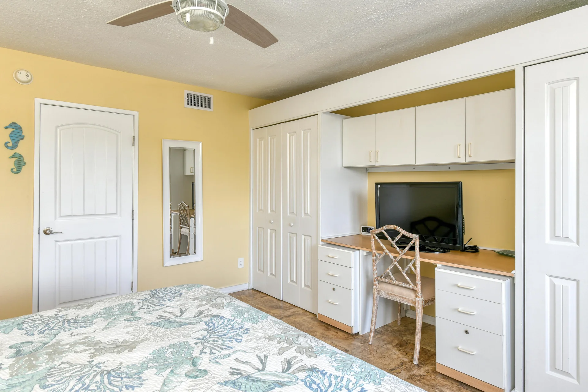 Built-in desk with white drawers and cabinets, a cushioned wooden chair, and a wall-mounted mirror in the bedroom.