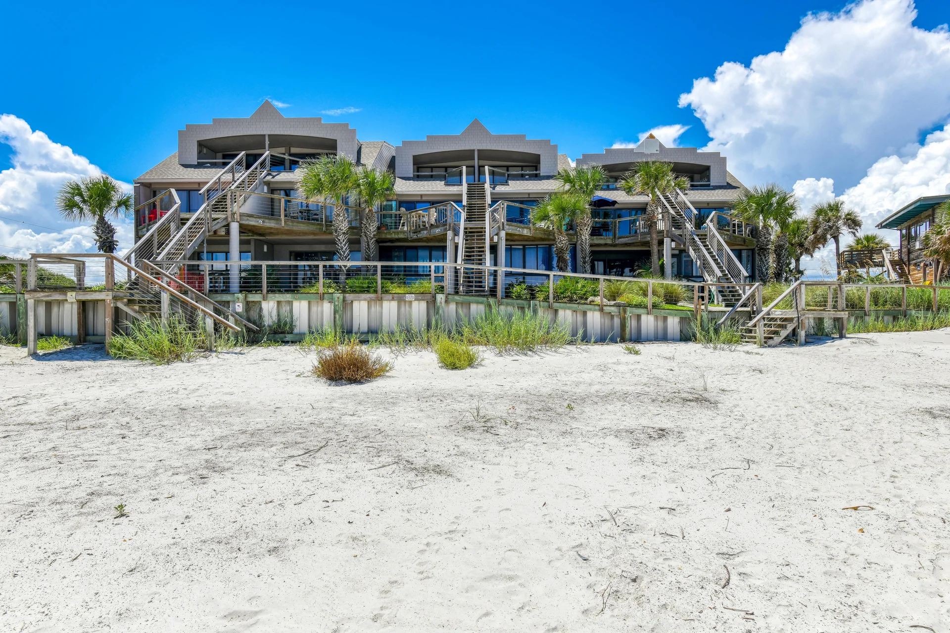 Front view of beachfront condos with wooden staircases, palm trees, and a wide sandy beach under a blue sky.