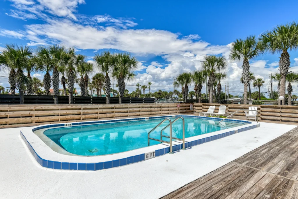 Rectangular pool with blue tile trim set in white concrete, surrounded by wood decking and palm trees under a blue sky.