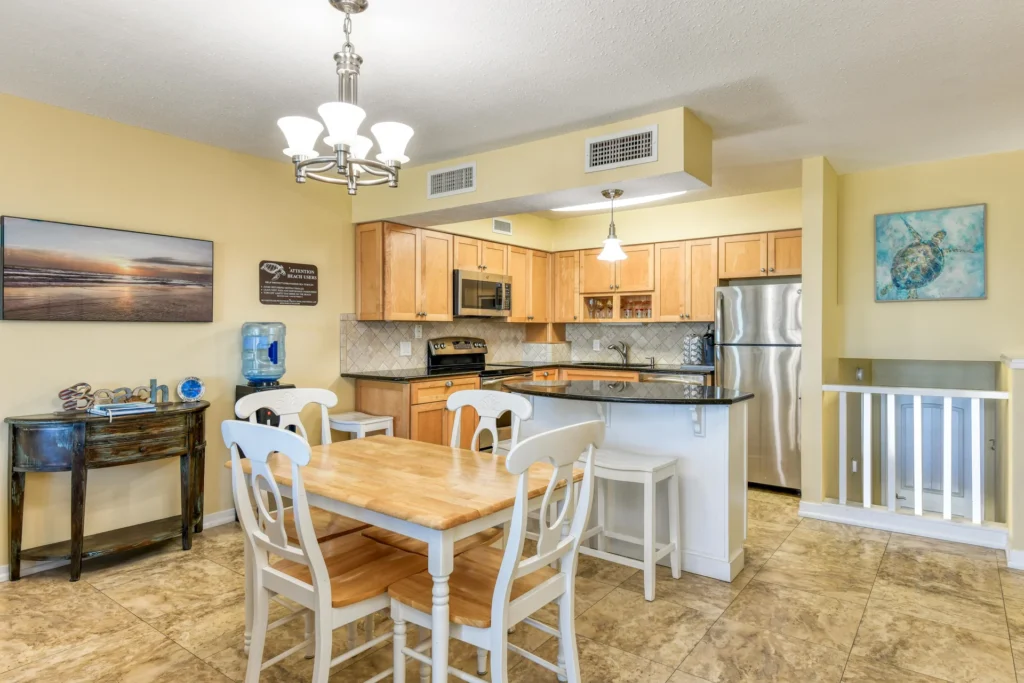 Dining area with wooden table and white chairs beside an open kitchen featuring light wood cabinets and granite counters.