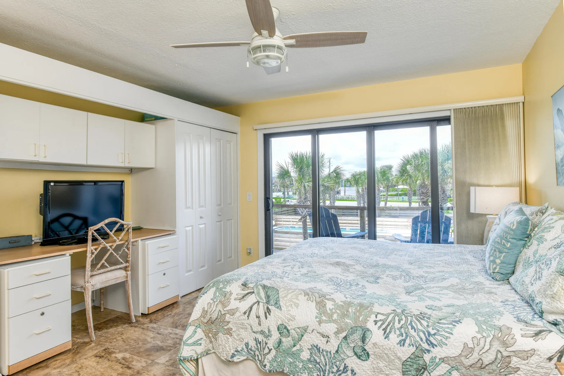 Bedroom with patterned coastal quilt on the bed, built-in desk with TV, and sliding glass doors opening to a balcony.