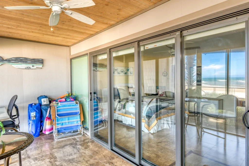 Covered balcony with tiled floor, ceiling fan, stacked beach gear, and sliding glass doors reflecting ocean views.