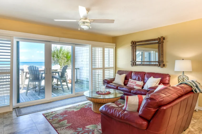 Living room with red leather sectional, glass-top wicker coffee table, floral rug, and ocean-view sliding doors.