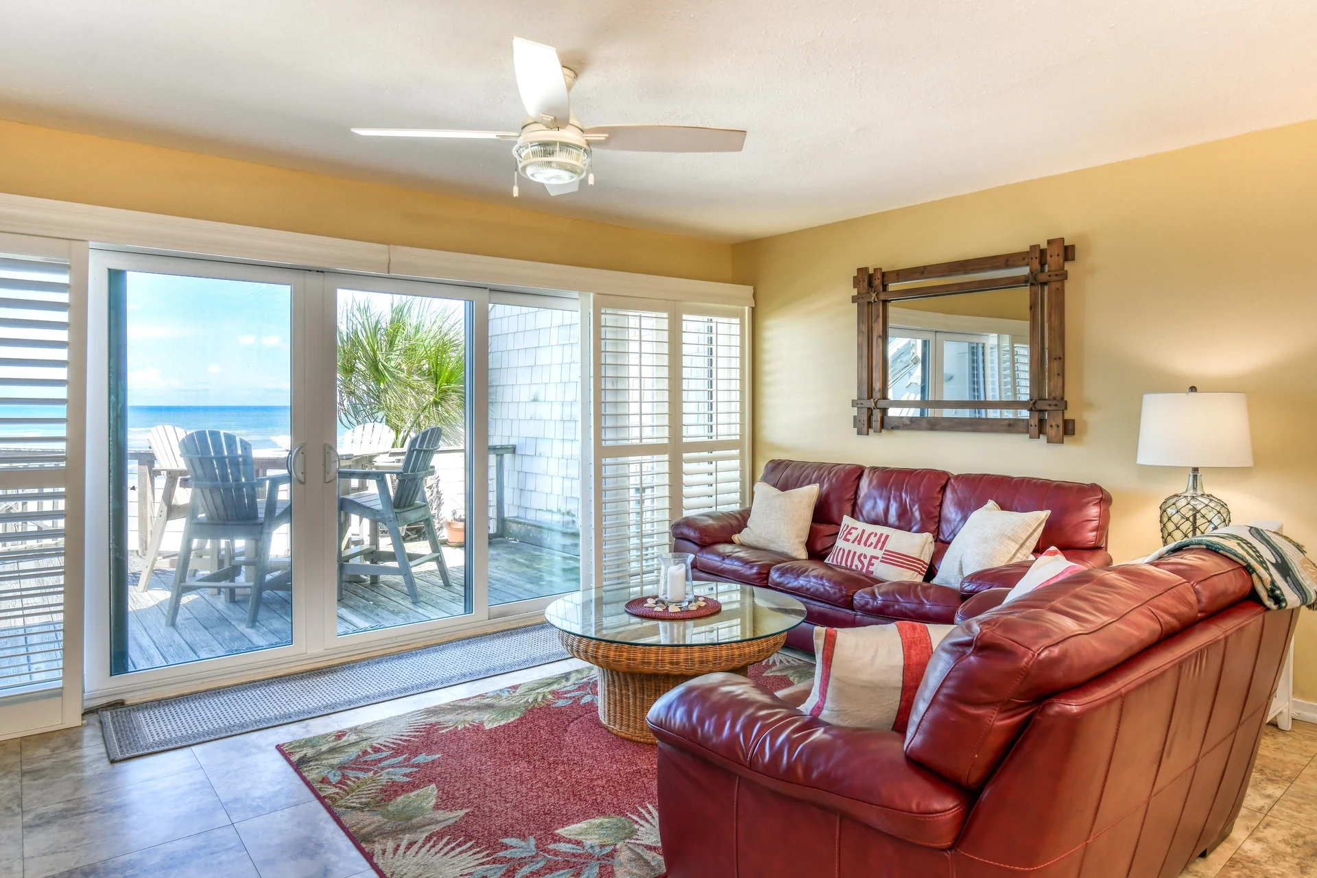 Living room with red leather sectional, glass-top wicker coffee table, floral rug, and ocean-view sliding doors.