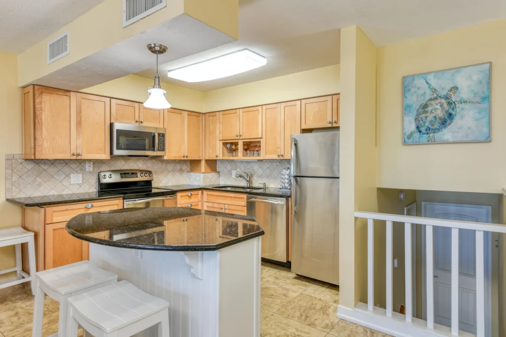 Kitchen features light wood cabinets, black granite countertops, stainless steel appliances, and a small breakfast bar with white stools.